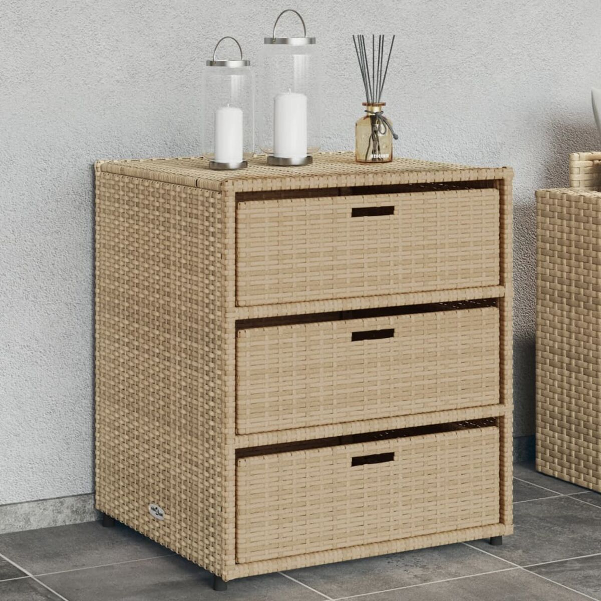 Ein beigefarbenes Rattan-Schränkchen mit drei Schubladen, gekrönt von Kerzen und einem Duftzerstäuber, vor einer grauen Wand.