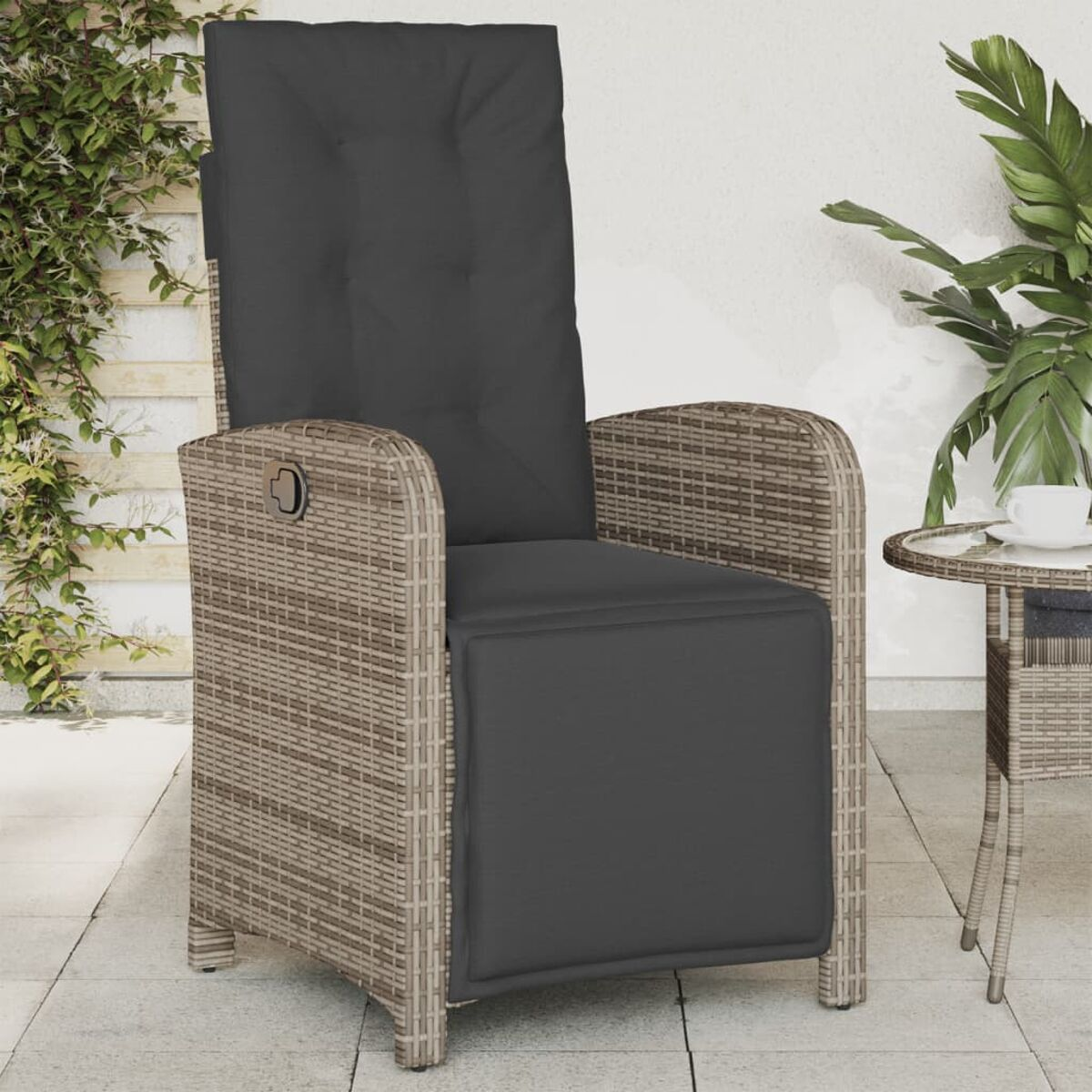 Rattan-Liegestuhl mit grauem Kissen auf einer Terrasse. Ein kleiner Tisch und eine Pflanze sind im Hintergrund.