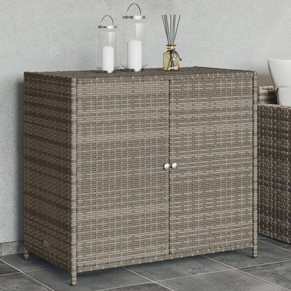 Grauer Rattan-Schrank mit zwei Türen. Zwei Kerzen und eine Vase stehen oben. Auf Fliesenboden.