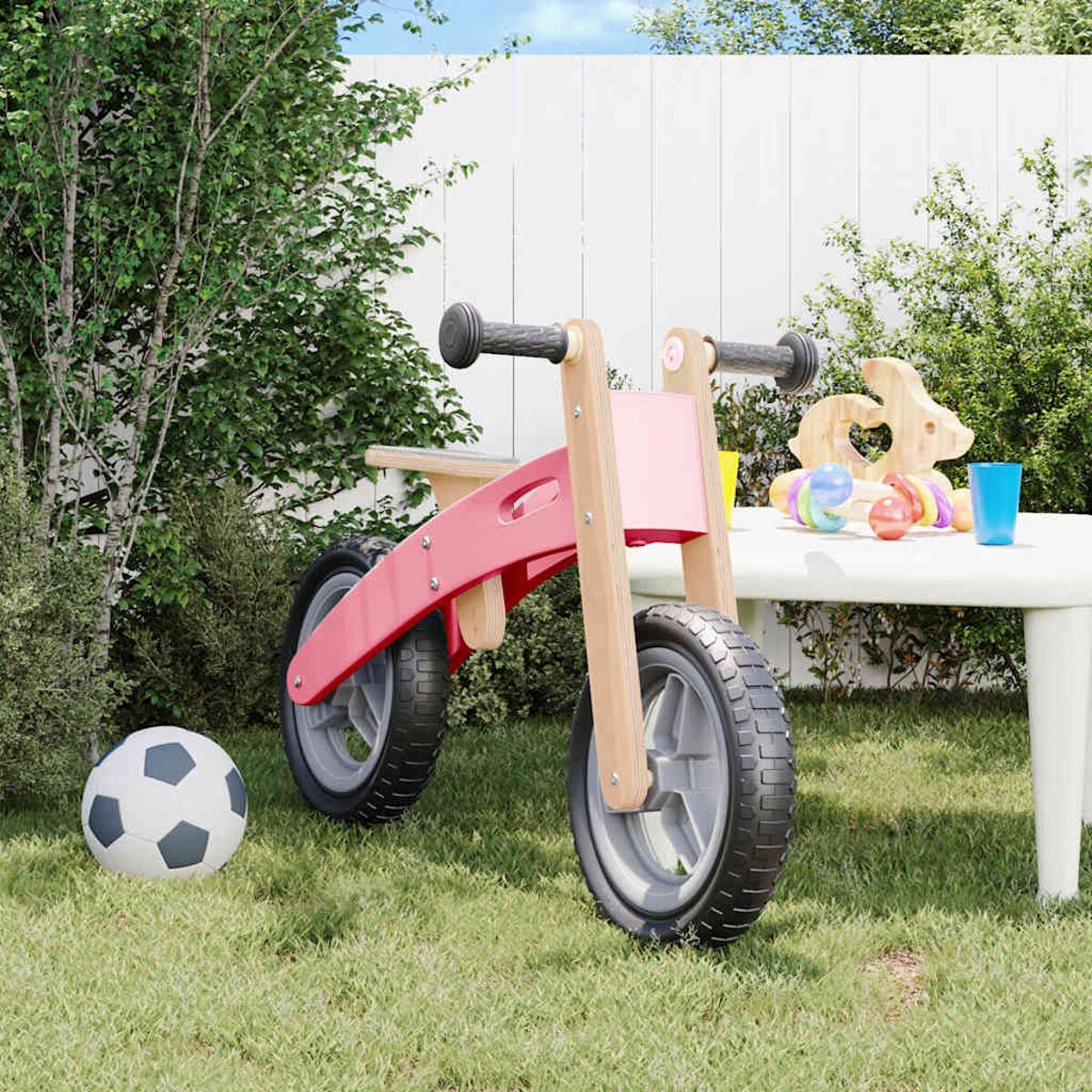 Ein rotes und hölzernes Laufrad, ein Fußball und Spielzeug in einer grasbewachsenen Fläche mit weißem Zaun.