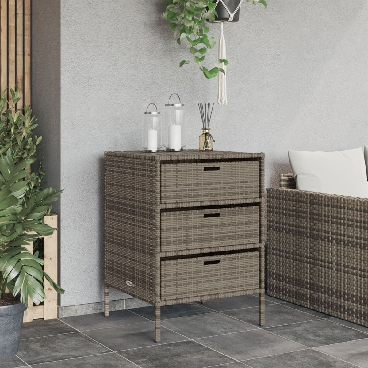 Rattanschränkchen mit drei Schubladen und zwei Kerzen. Pflanze und Sofa an den Seiten. Grauer Boden.