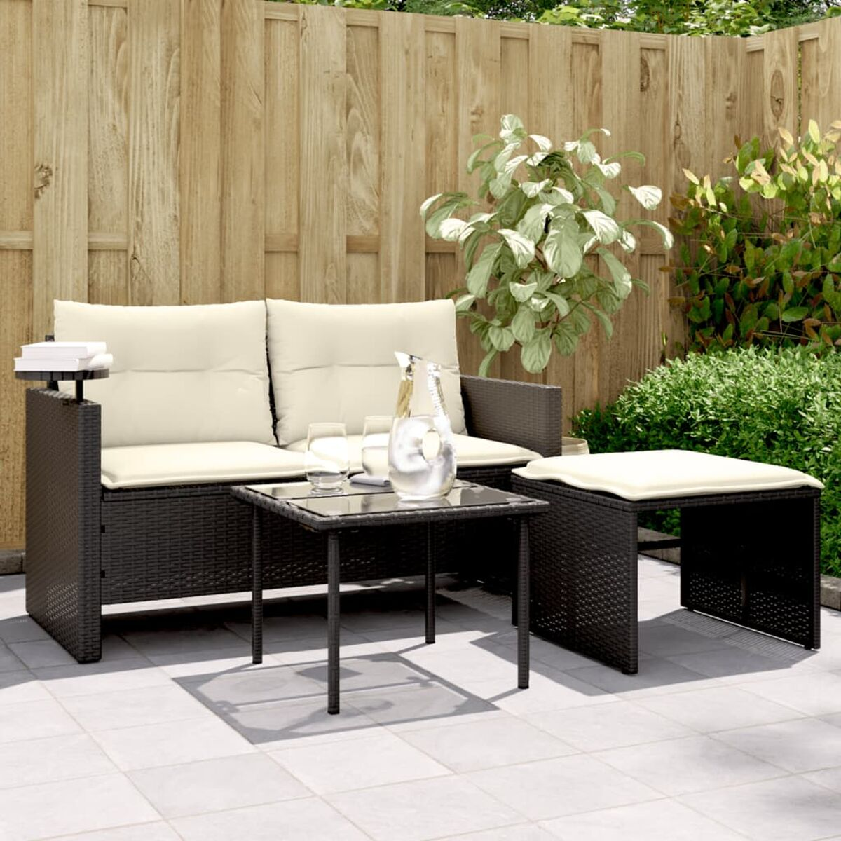 Set da patio: un divano, un tavolino e uno sgabello. Tutti in vimini nero. Recinzione in legno dietro.