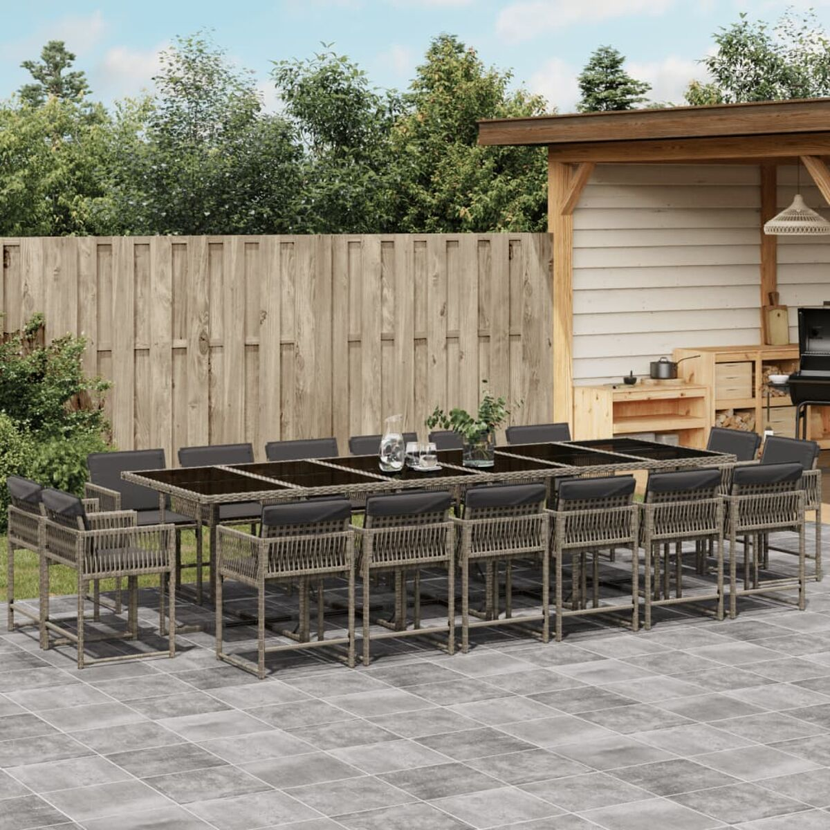 Set da pranzo per esterni con un lungo tavolo e sedie con cuscini grigi. Include una pergola e una cucina esterna.