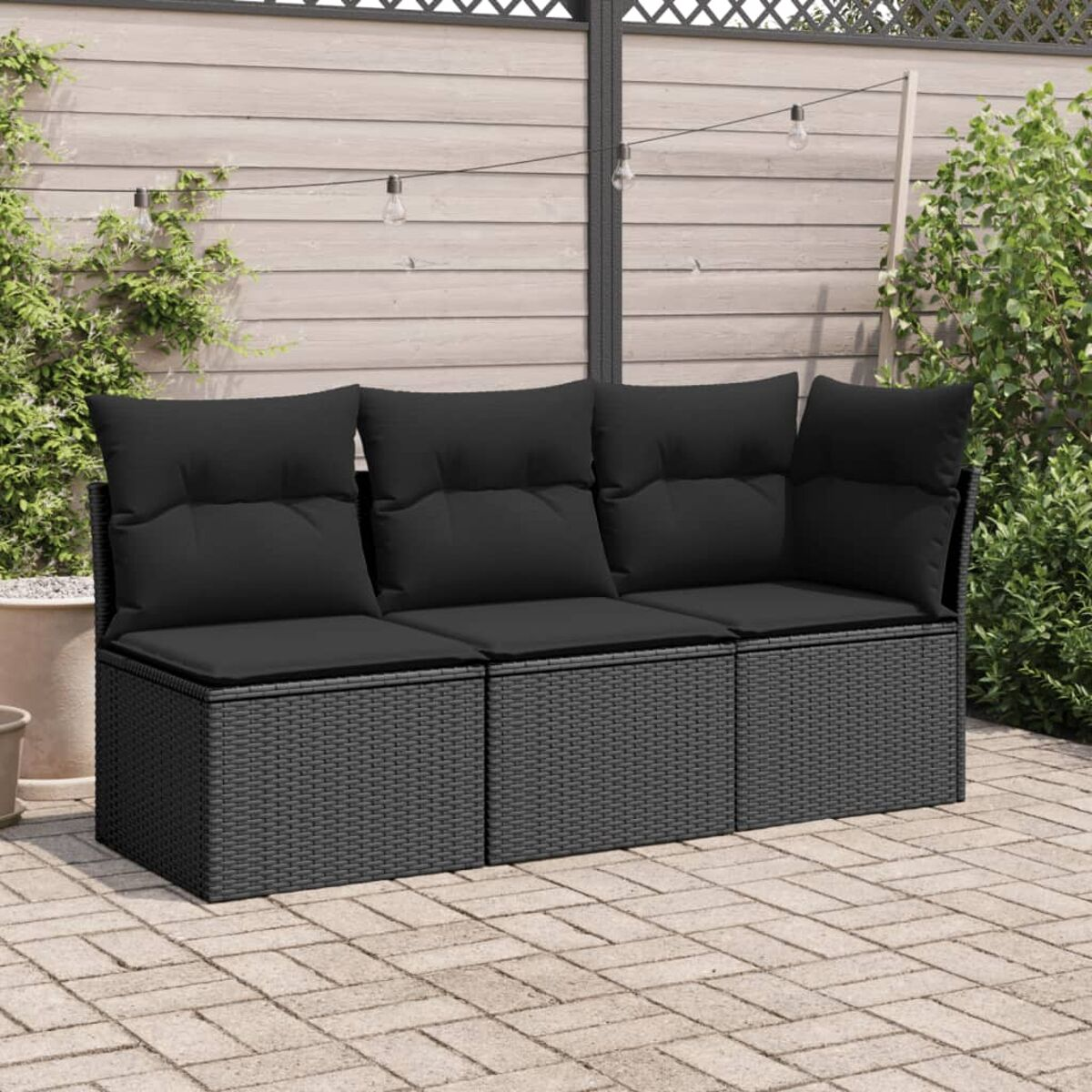 Schwarzes Outdoor-Sofa aus Korbgeflecht mit schwarzen Kissen. Ziegelterrasse, Holzzaun und Lichterketten.