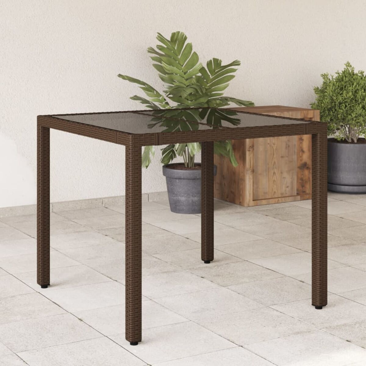 Quadratischer brauner Outdoor-Tisch aus Korbgeflecht mit Glasplatte. Pflanzen im Hintergrund.