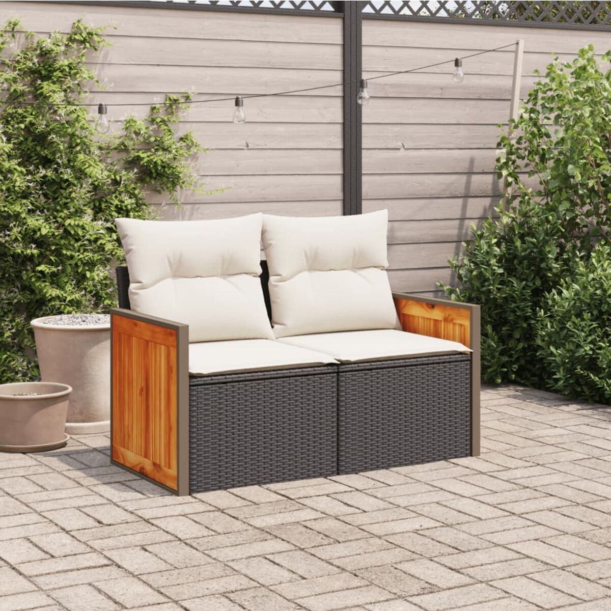 Gartensofa mit grauem Geflecht, Holzseiten, weißen Kissen und einer Backsteinterrasse. Grüne Pflanzen im Hintergrund.