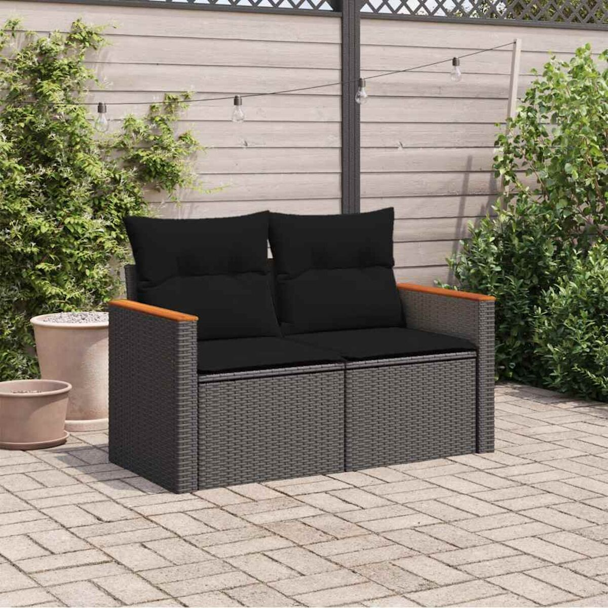 Outdoor-Liebesitz mit grauem Geflecht, Holzarmlehnen und schwarzen Kissen. Pflanzen und Pflastersteine im Hintergrund.