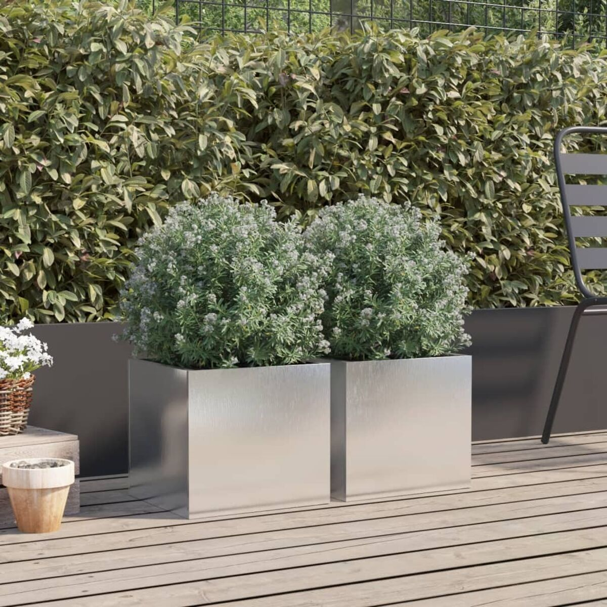 Zwei quadratische, metallische Pflanzgefäße mit grünen Pflanzen, auf einem Holzdeck, mit Heckenhintergrund.