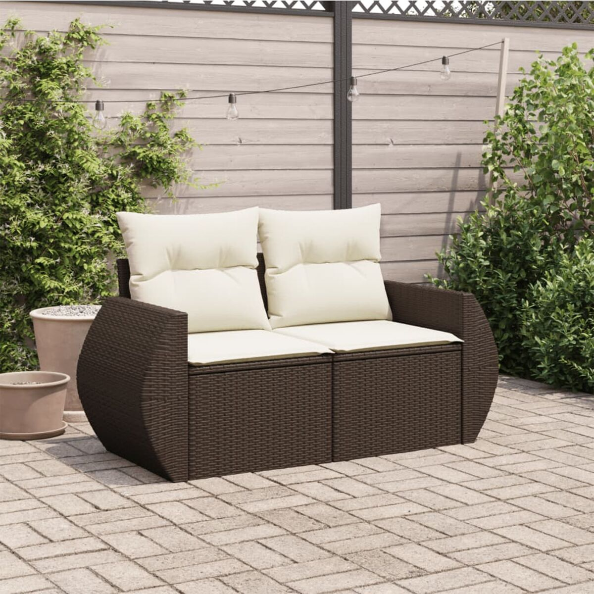 Divano da esterno in rattan marrone con cuscini color crema. È su un patio in pietra con sfondo verde.