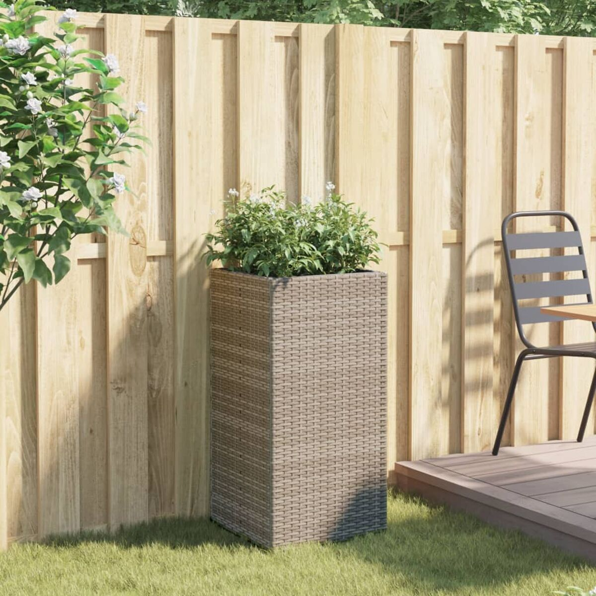 Fioriera alta in rattan con vegetazione, sull'erba, vicino a una recinzione.