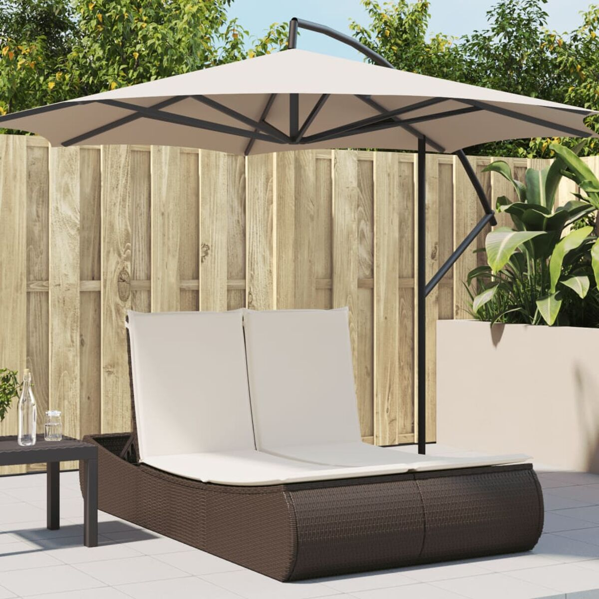 Lettino prendisole in vimini marrone a due posti con cuscini beige e ombrellone. Staccionata in legno sullo sfondo.