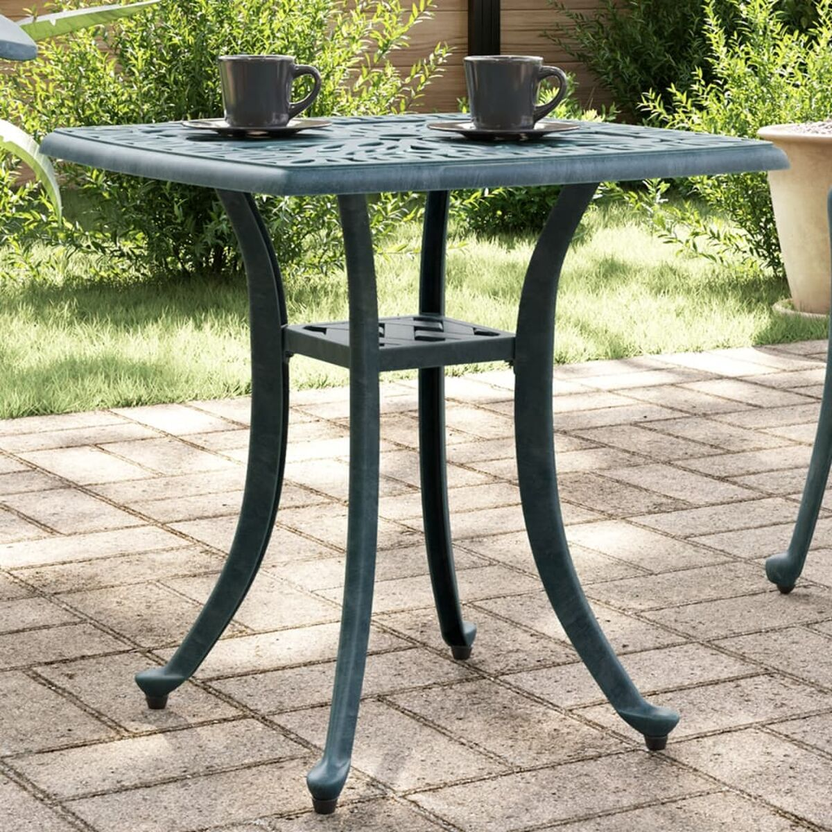 Quadratischer, dunkel türkisfarbener Terrassentisch mit zwei Tassen, umgeben von Grün.