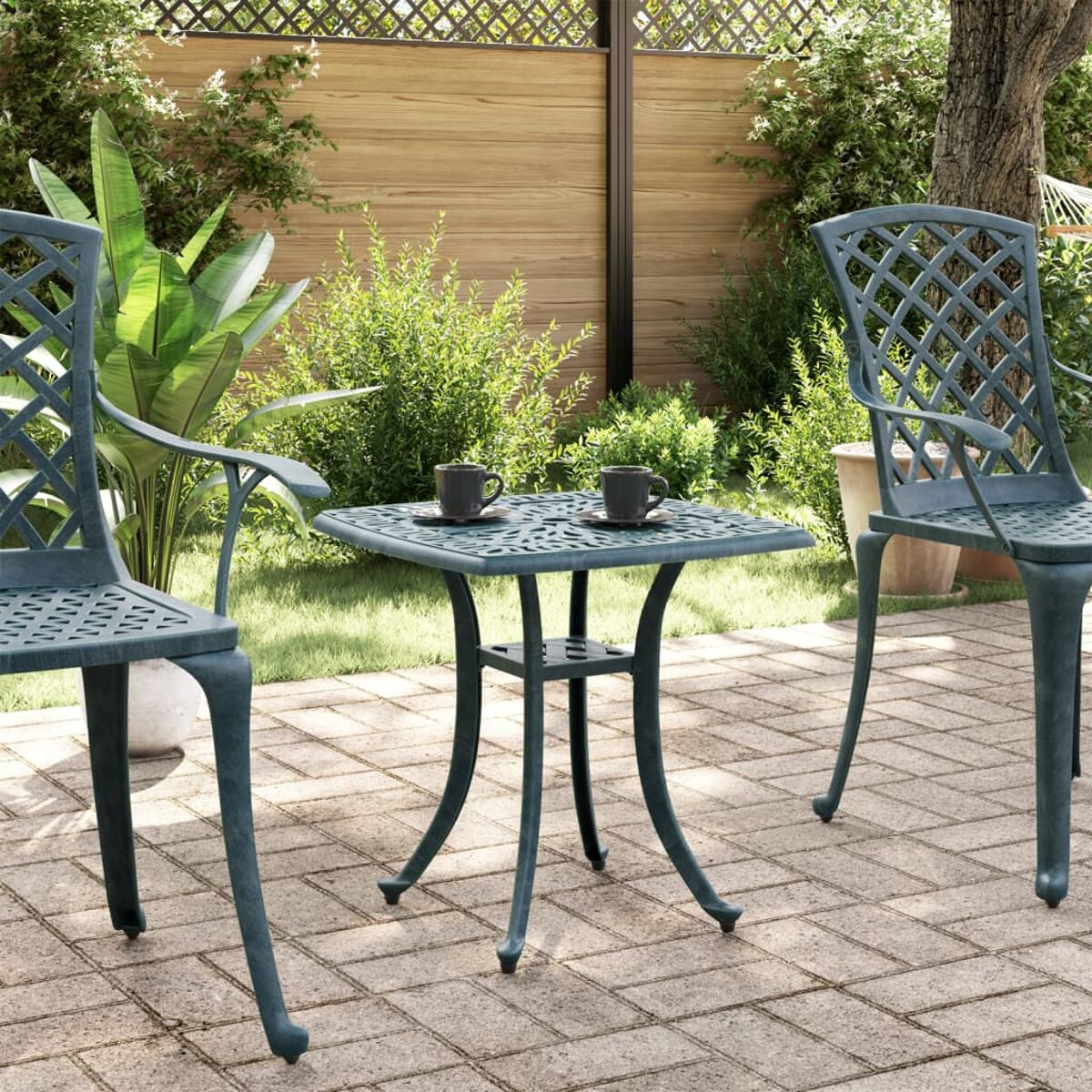 Zwei tealfarbene Metallstühle und ein Tisch auf einer Terrasse mit zwei Tassen. Pflanzen und Holzzaun im Hintergrund.