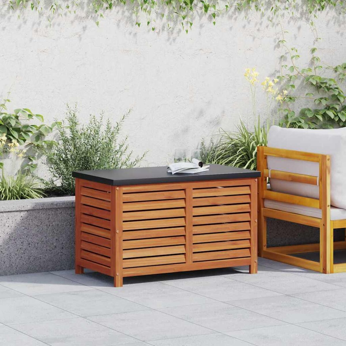 Holz-Aufbewahrungsbox mit schwarzer Oberseite, einem Glas und einem Buch auf einer gefliesten Terrasse, vor einer Wand und Pflanzen.