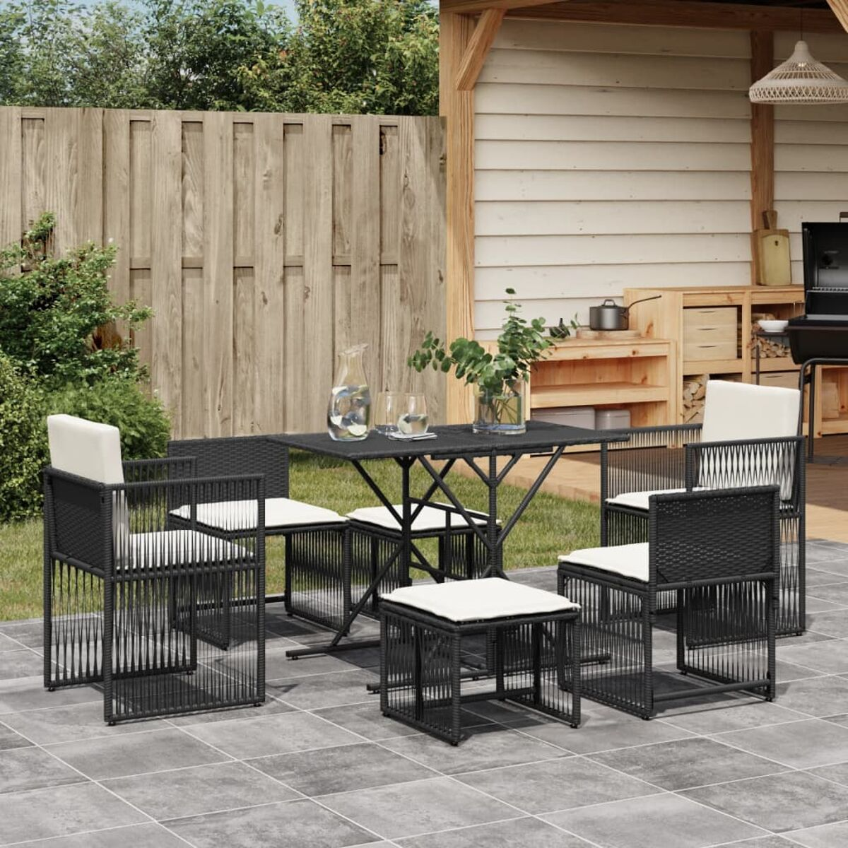 Essgruppe im Freien. Schwarze Stühle, Tisch und Kissen auf einer Terrasse. Hintergrund umfaßt einen Zaun und Schuppen.