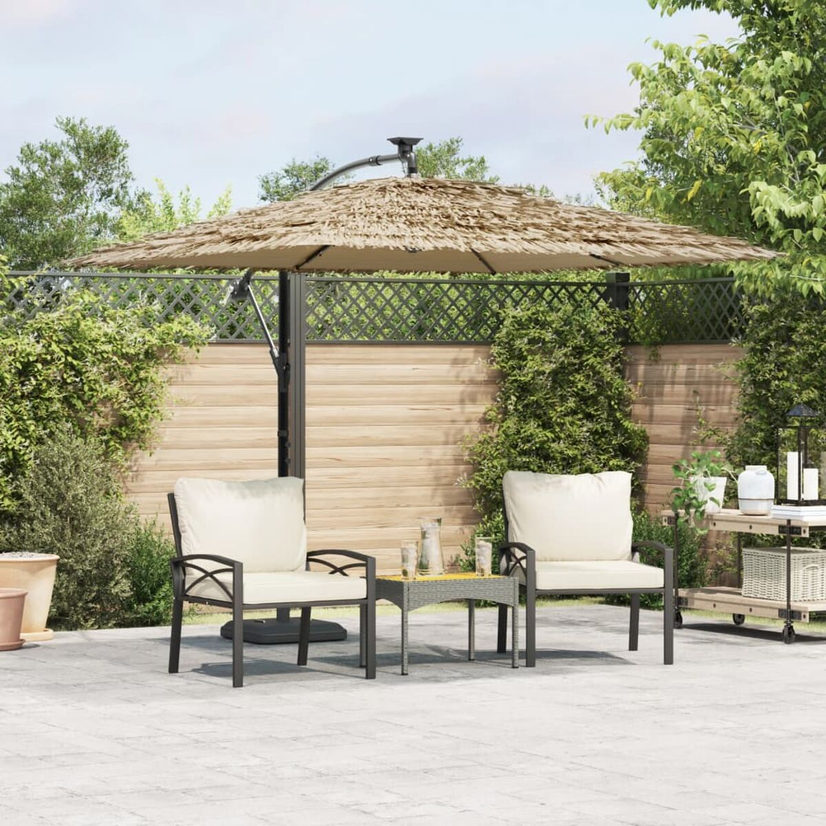 Terrassenset mit Schirm, zwei Stühlen und Tisch. Beiger Schirm, schwarzer Rahmen. Außenbereich mit Grün.