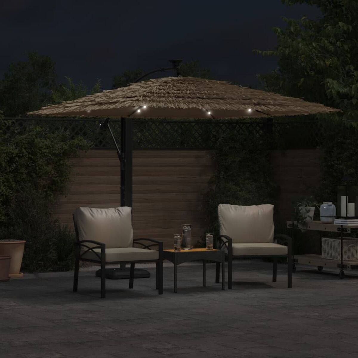 Terrassenmöbel bei Nacht. Zwei Stühle, ein Tisch und ein Schirm mit Lichtern, umgeben von Grün.