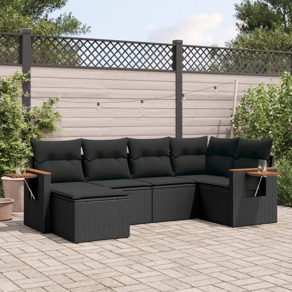 Schwarze Outdoor-Eckcouch aus Korbgeflecht mit schwarzen Kissen, Holzarmlehnen, Tisch. Lichter und Zaun.
