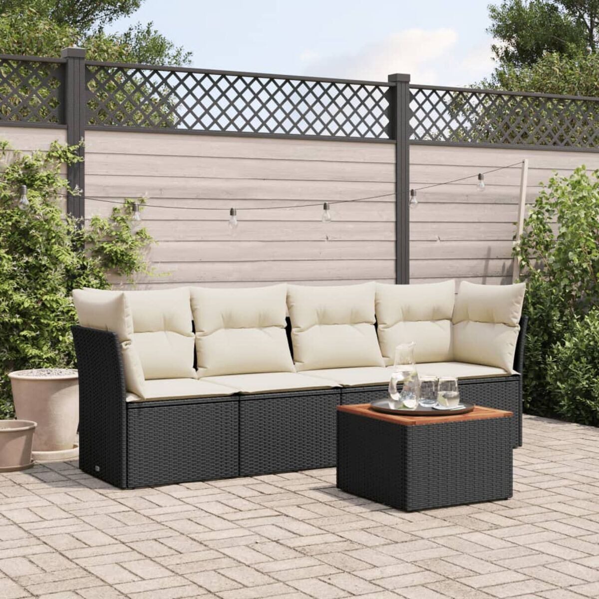 Schwarzes Korbmöbel-Set mit cremefarbenen Kissen, Tisch, Getränken und Gartenhintergrund.