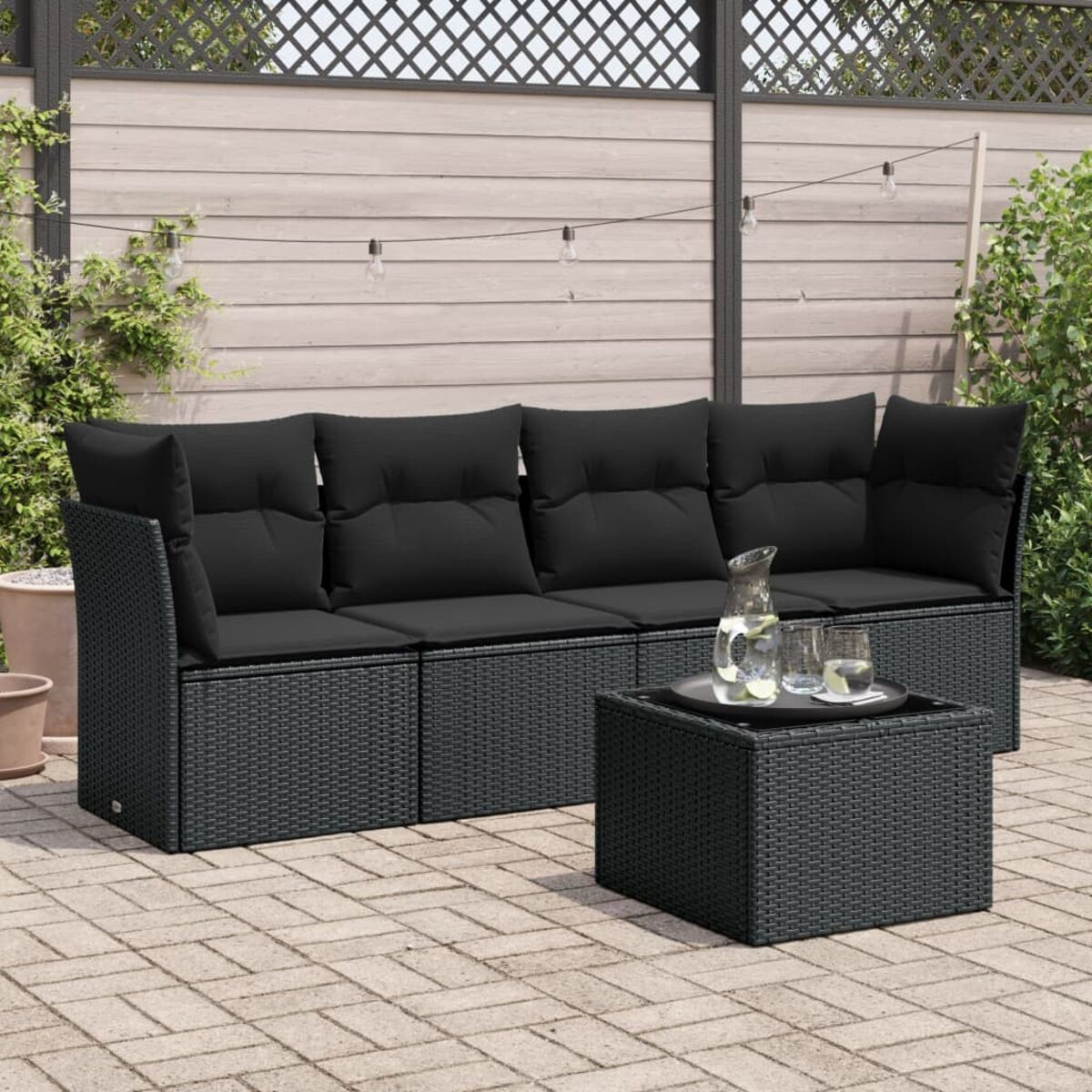 Schwarzes Outdoor-Wicker-Sofa mit schwarzen Kissen und einem Tisch mit Gläsern. Ziegelterrasse, Zaun im Hintergrund.