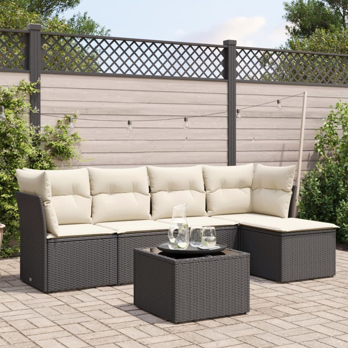 Outdoor-Ecksofa mit beigen Kissen und schwarzem Korbboden, und Tisch. Gepflasterte Terrasse mit Grün.