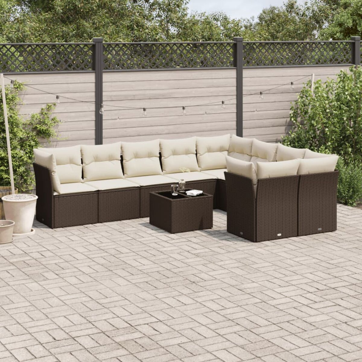Divano componibile da esterno marrone scuro con cuscini color crema, un tavolino e un patio in mattoni. Verde e recinzione sullo sfondo.