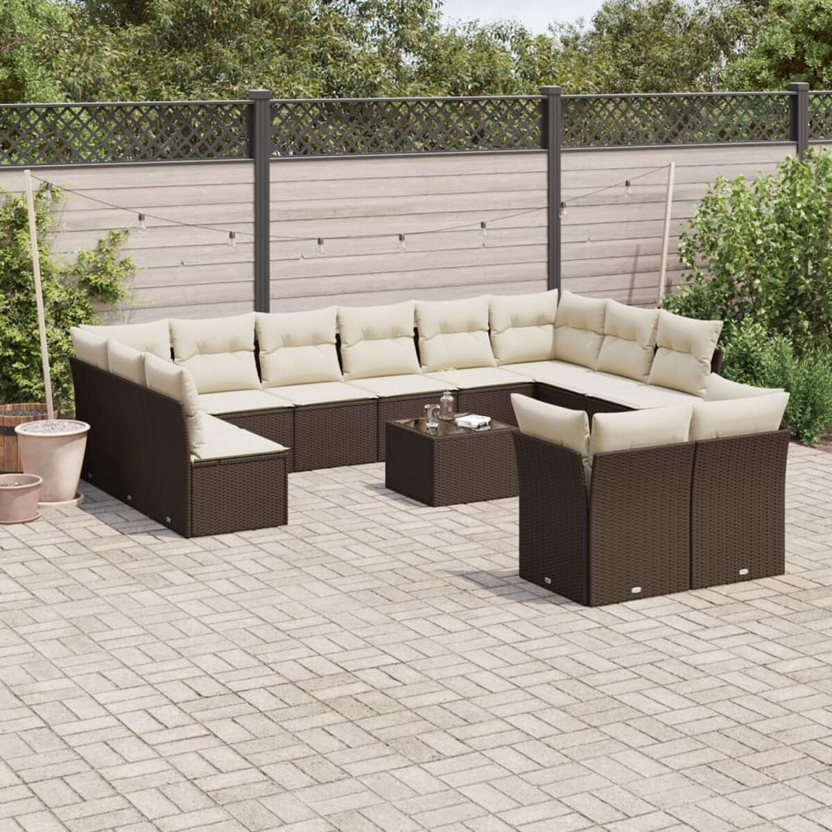Divano da esterno in rattan marrone con cuscini color crema, tavolino e patio in mattoni, con recinzione sullo sfondo.