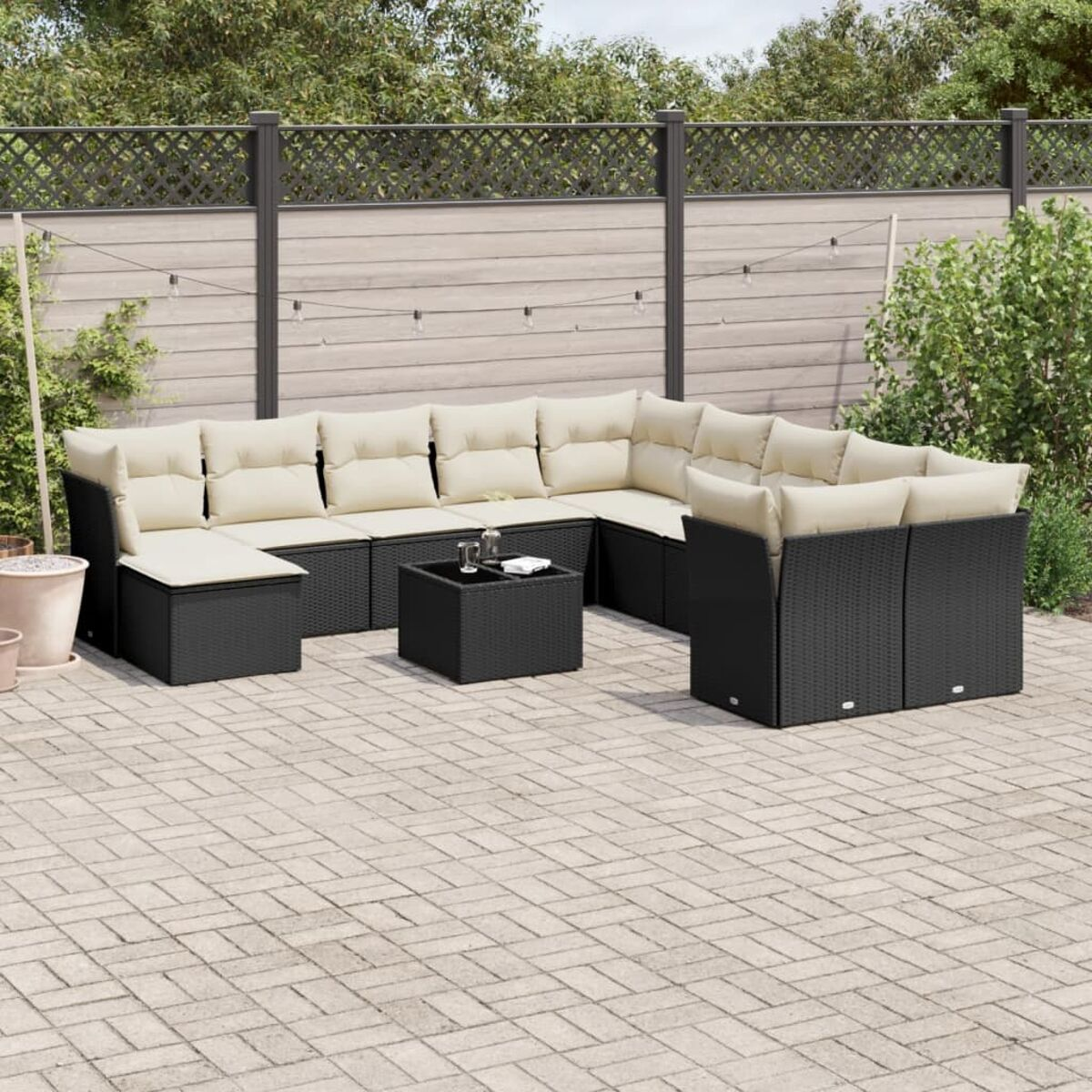 Outdoor-Eckgarnitur. Sie ist schwarz mit cremefarbenen Kissen. Sie steht auf einer Terrasse mit Zaun und Pflanzen.
