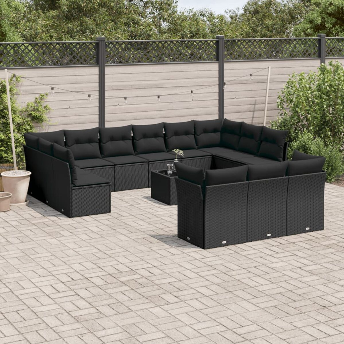 Schwarzes Outdoor-Ecksofa-Set mit schwarzen Kissen und Couchtisch. Auf einer Backstein-Terrasse. Grünes Laub im Hintergrund.