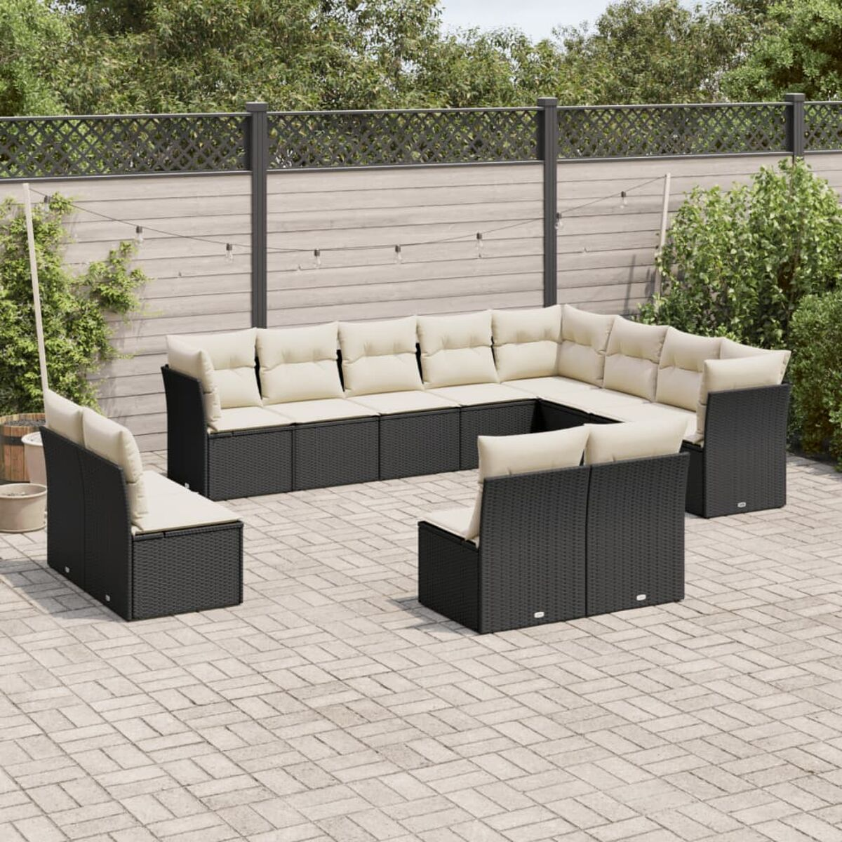 Schwarze Outdoor-Eckcouch aus Korbgeflecht mit beigefarbenen Kissen auf einer gepflasterten Terrasse. Ein Zaun und Grün im Hintergrund.