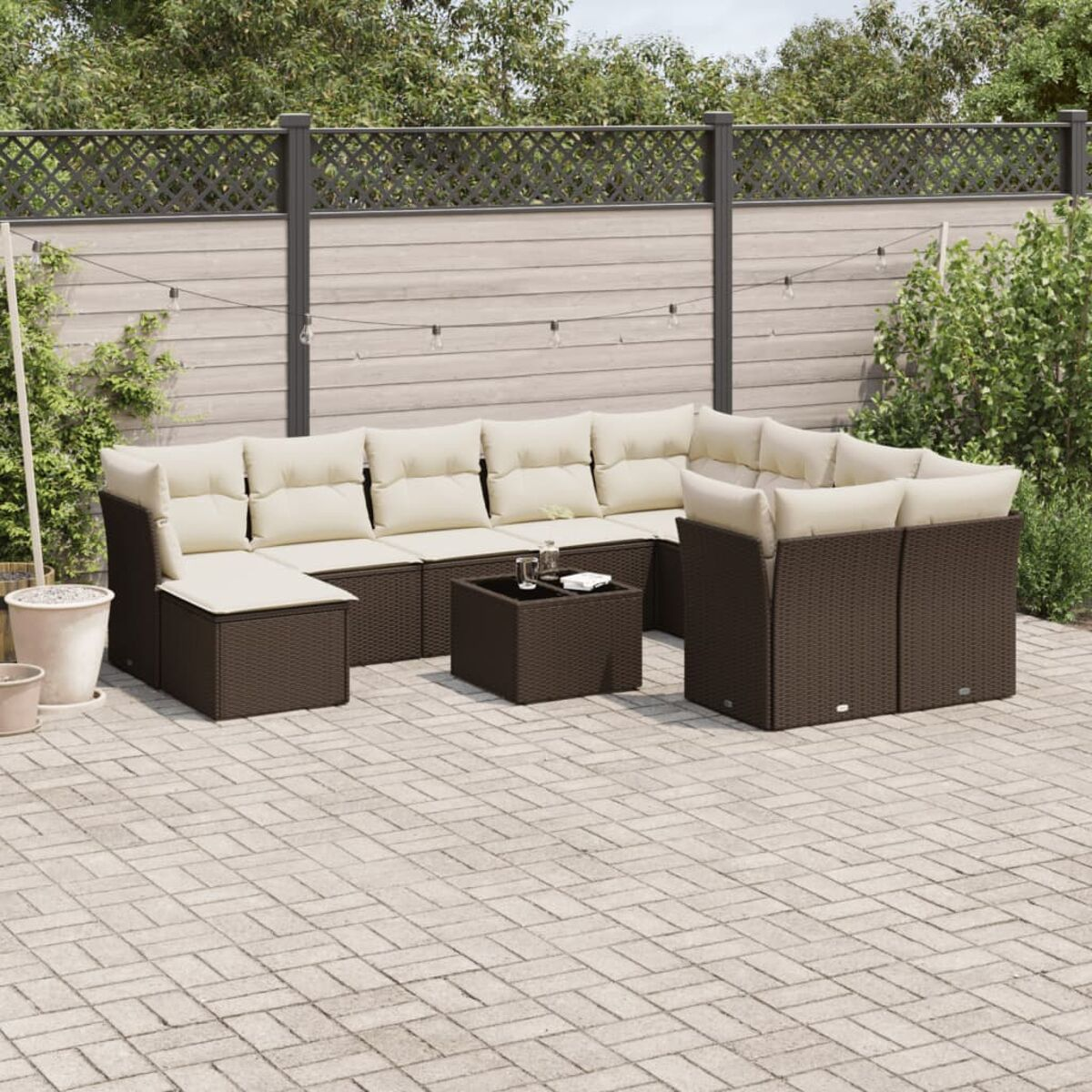 Set divano da esterno in vimini marrone con cuscini color crema, tavolo quadrato e piante in giardino.