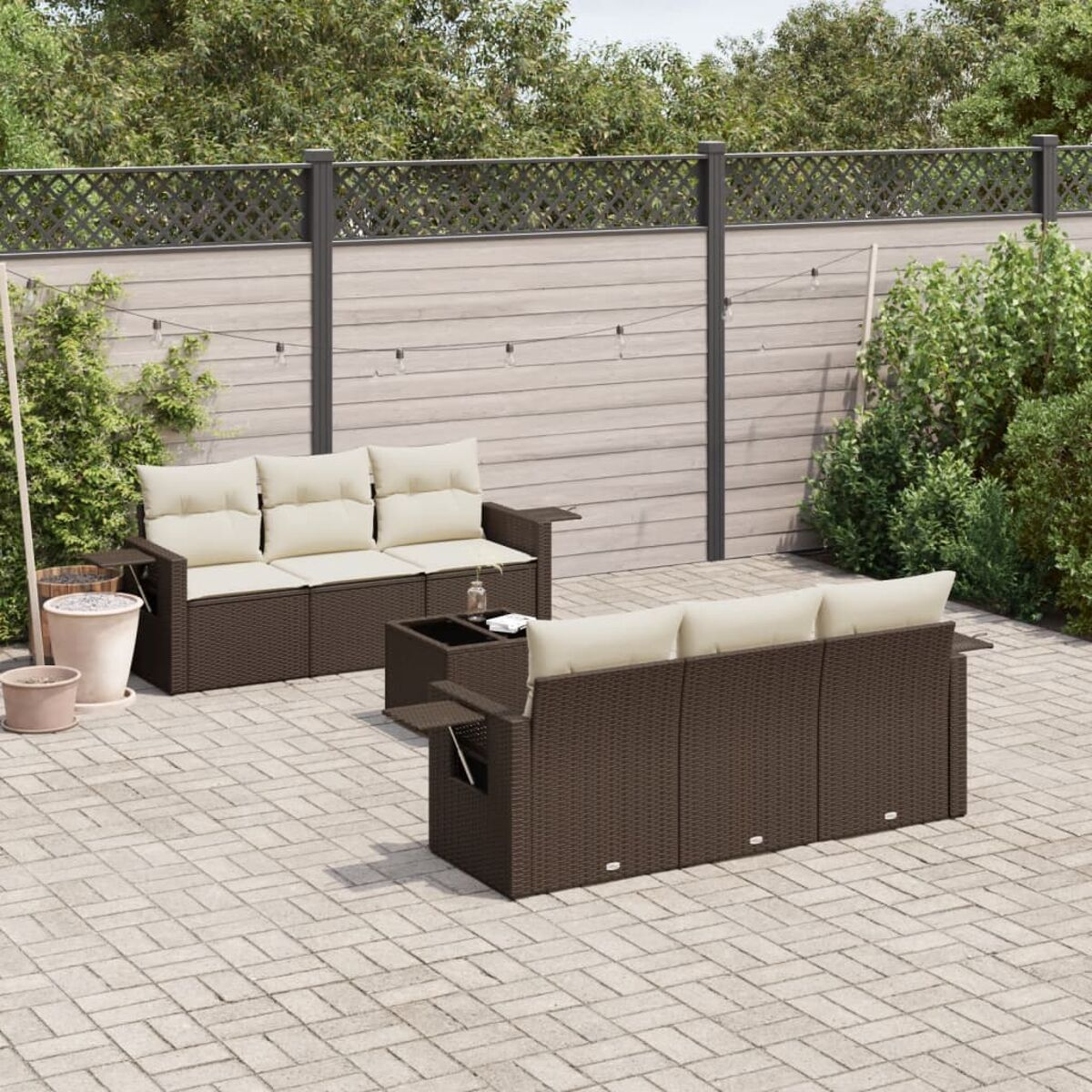Set divano da esterno in vimini marrone con cuscini beige. Patio con pavimentazione in mattoni grigi. Piante verdi e recinzione sullo sfondo.