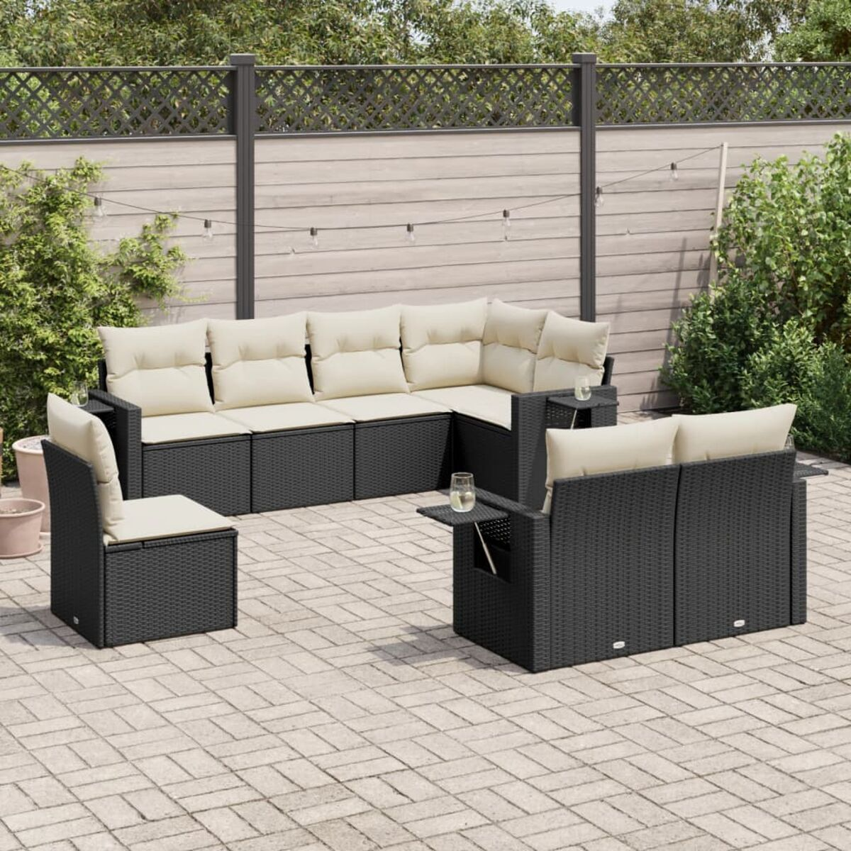 Außenbereich mit schwarzen Rattanmöbeln, cremefarbenen Kissen und gepflasterter Terrasse. Pflanzen und Zaun sichtbar.