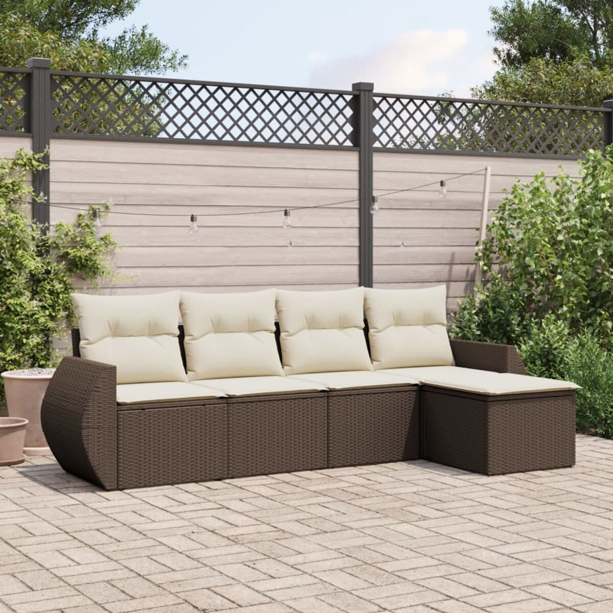 Set divano in vimini marrone con cuscini beige. Ambiente esterno su patio in mattoni. Recinzione e piante dietro.