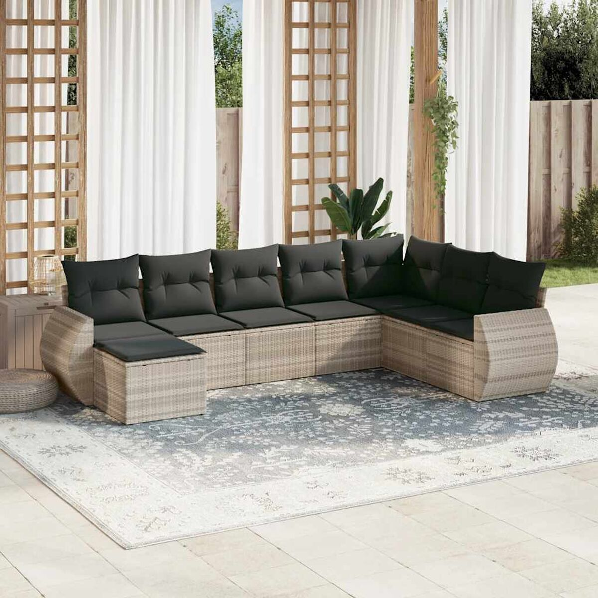 Rattan-Sofa-Set für draußen mit dunklen Kissen auf einem gemusterten Teppich, neben weißen Vorhängen auf einer Terrasse.