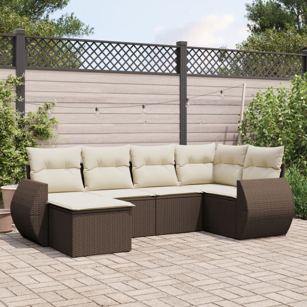 Ein braunes Rattan-Ecksofa mit cremefarbenen Kissen auf einer Terrasse. Ein Holzzaun und Grün im Hintergrund.