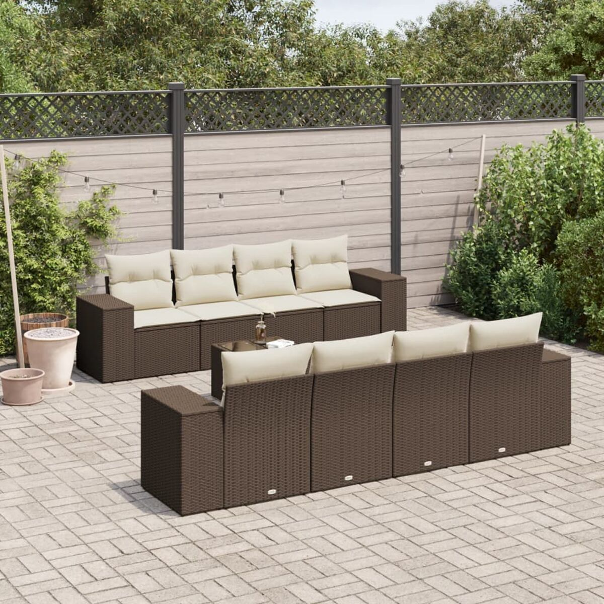 Set divano da esterno in vimini marrone con cuscini beige su un patio. Lo sfondo include recinzione e verde.