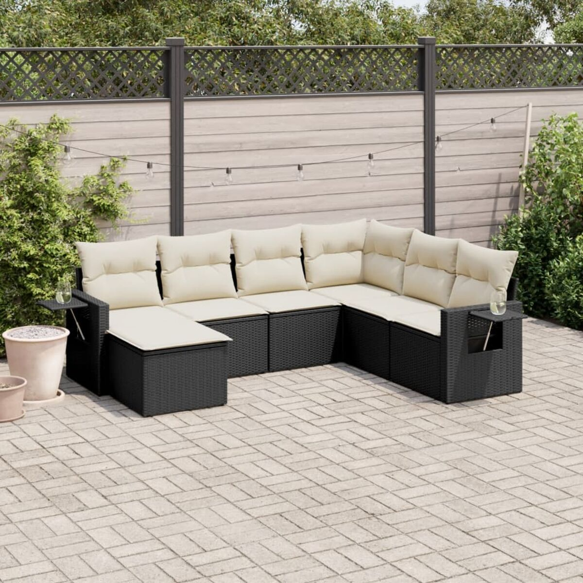 Schwarzes Ecksofa aus Korbgeflecht mit beigefarbenen Kissen. Auf einer Terrasse. Hintergrund: Zaun und Grün.