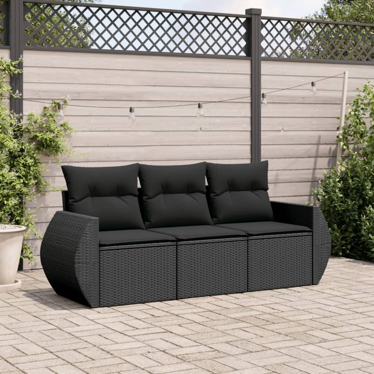 Schwarzes Korbmöbel mit schwarzen Kissen im Garten. Hintergrund: Zaun, Pflanzen, Lichterkette.