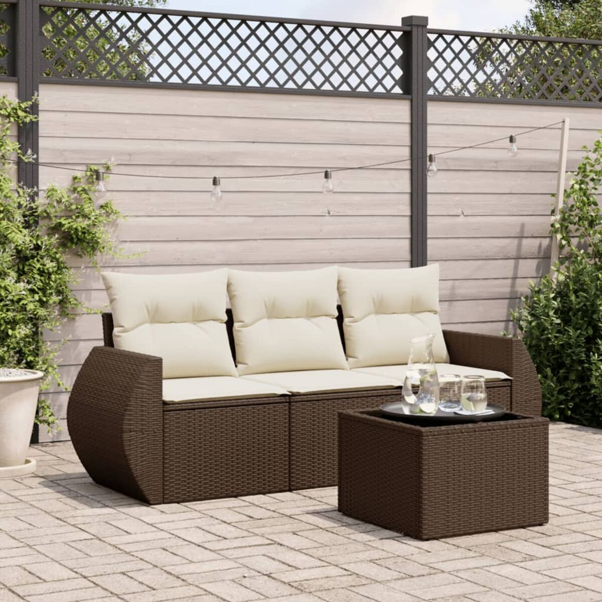 Divano in rattan marrone con cuscini color crema e tavolino. Lo sfondo include una recinzione e piante.