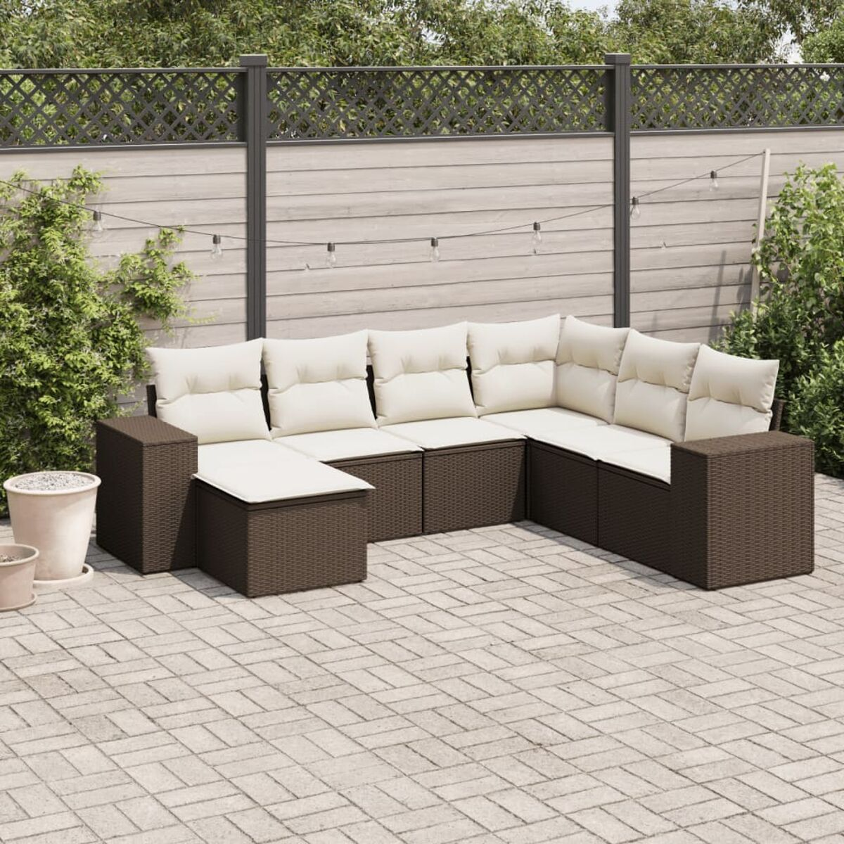 Divano angolare in vimini marrone con cuscini color crema su un patio. Una recinzione e piante sullo sfondo.