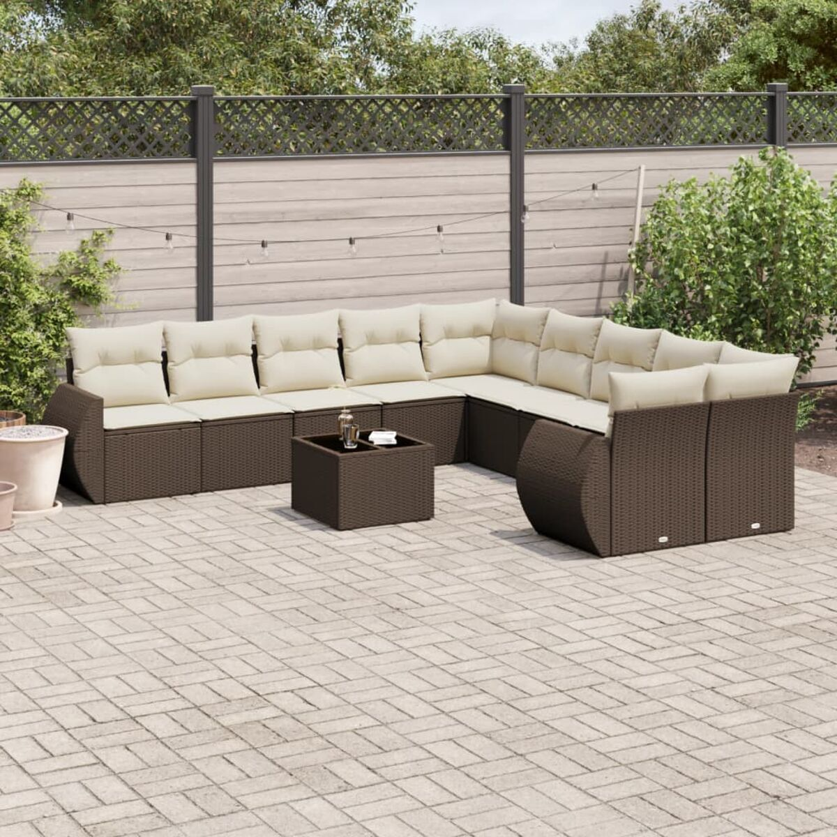 Divano componibile da esterno in vimini marrone. Cuscini beige. Tavolino quadrato. Patio in mattoni. Sfondo recintato.
