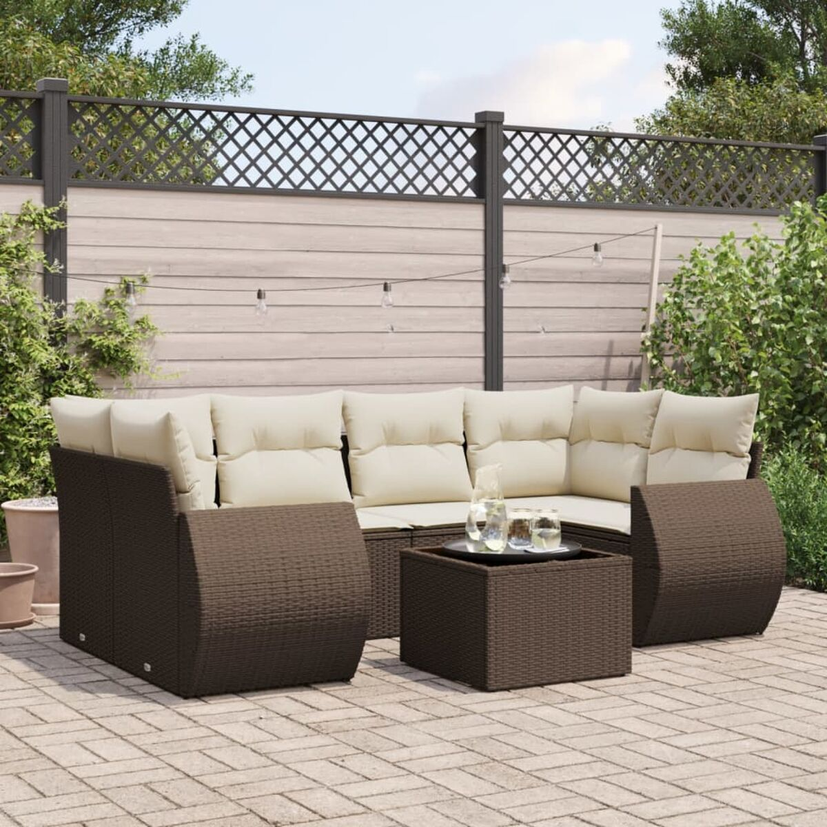 Divano componibile in rattan marrone con cuscini beige e tavolino quadrato. Recinzione, piante e luci dietro.
