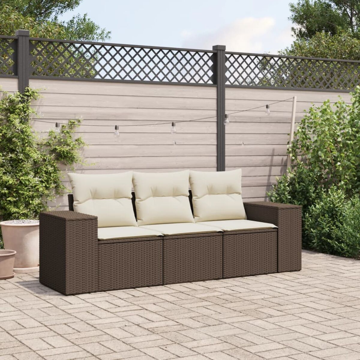 Divano in rattan marrone con cuscini color crema su un patio. Una recinzione in legno con luci e vegetazione sullo sfondo.