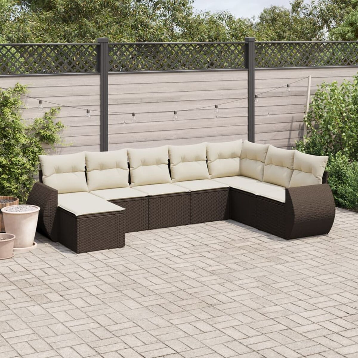 Divano componibile in vimini marrone con cuscini color crema, su un patio. Recinzione e verde sullo sfondo.