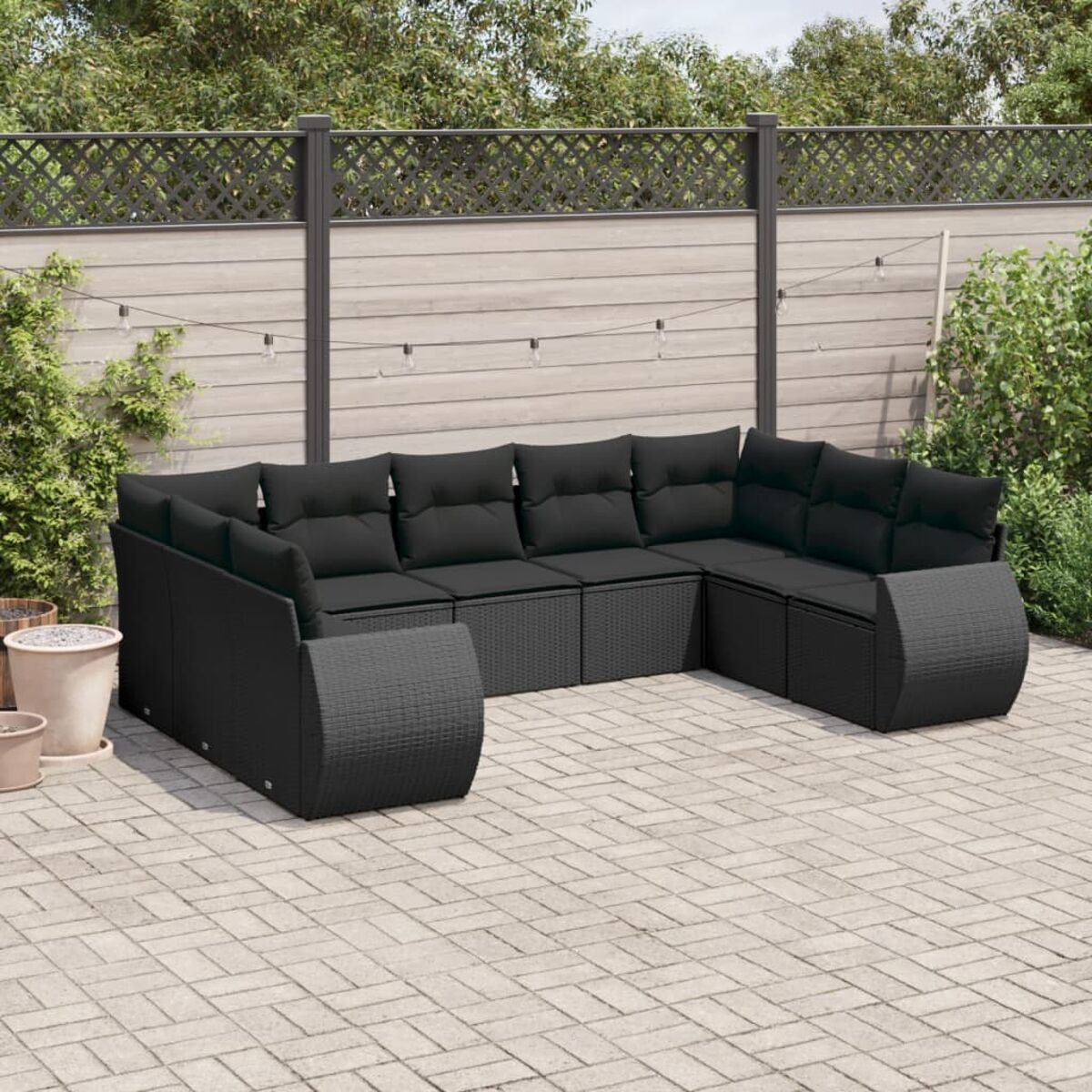 Outdoor-Terrassenset mit schwarzen Rattan-Sofas, Kissen und Ziegelboden. Holzzaun und grüner Hintergrund.