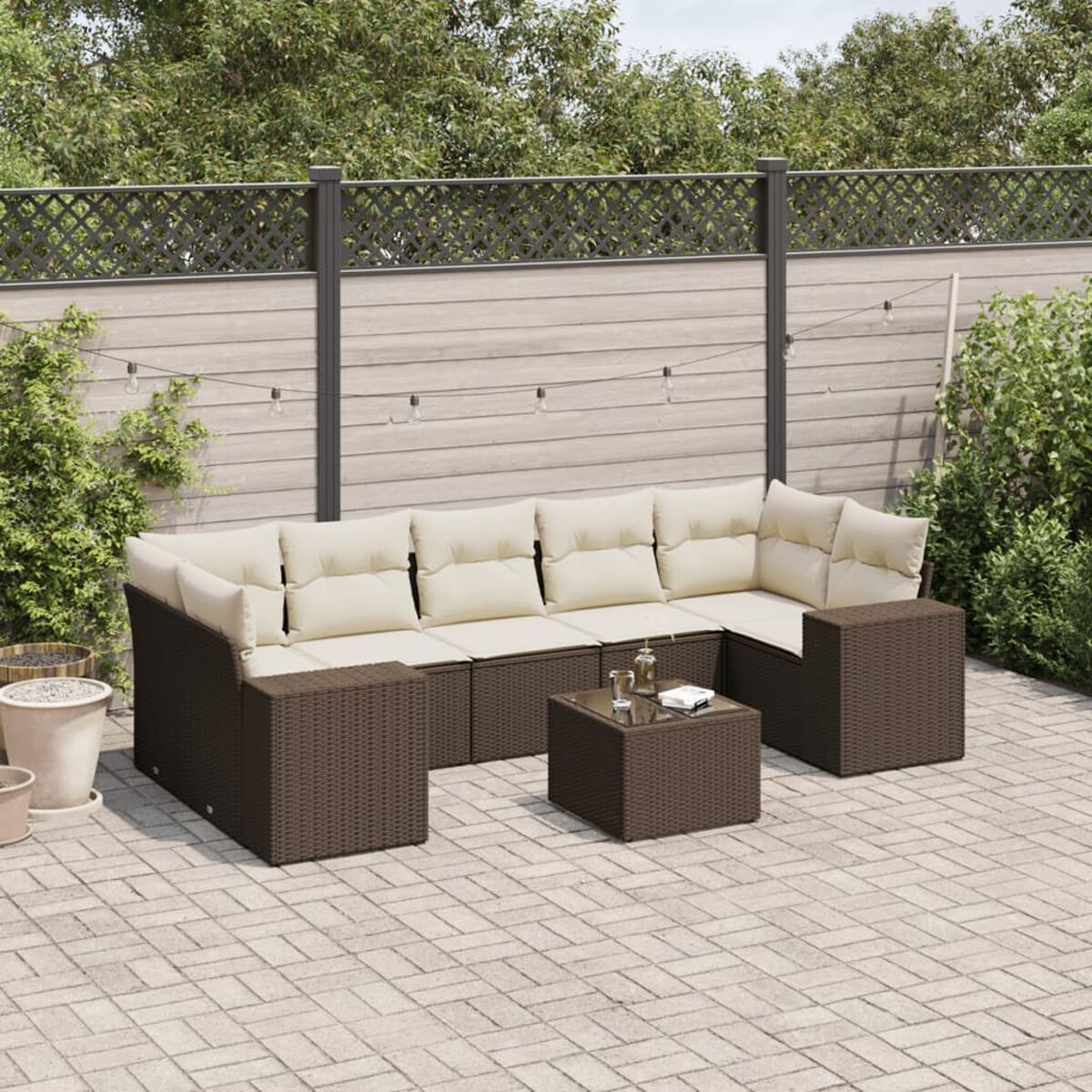 Set divano da esterno in rattan marrone con cuscini color crema e un tavolino su un patio in mattoni, con una recinzione in legno.