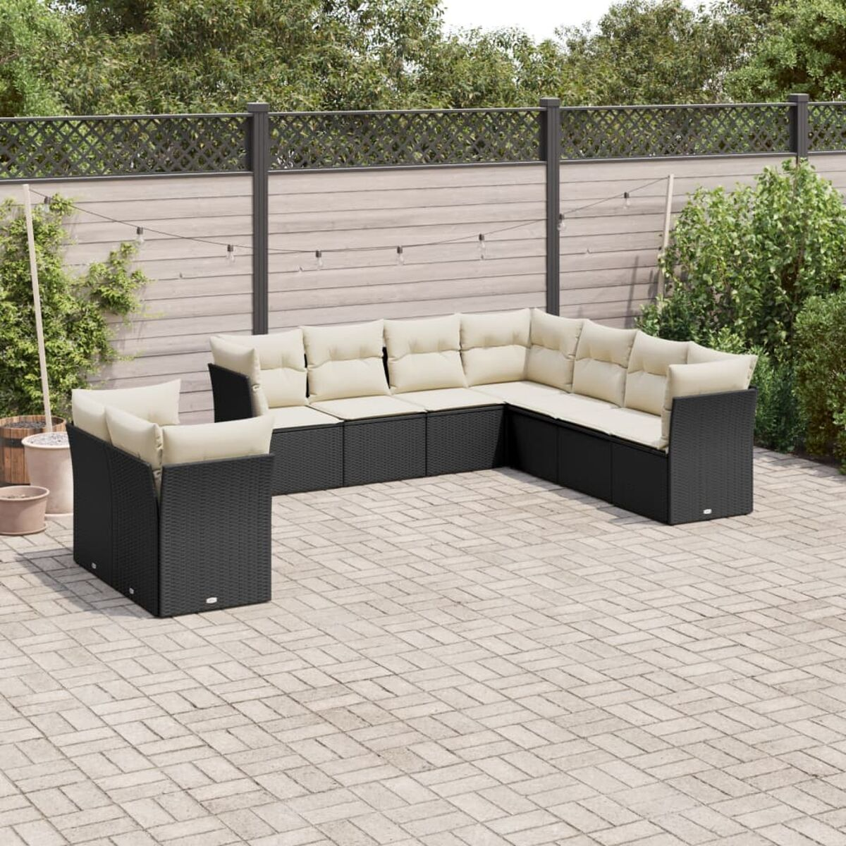 Schwarze Outdoor-Geflecht-Eckgarnitur mit beigen Kissen, Tisch und Getränken. Backstein-Terrasse, Zaun und Bäume.
