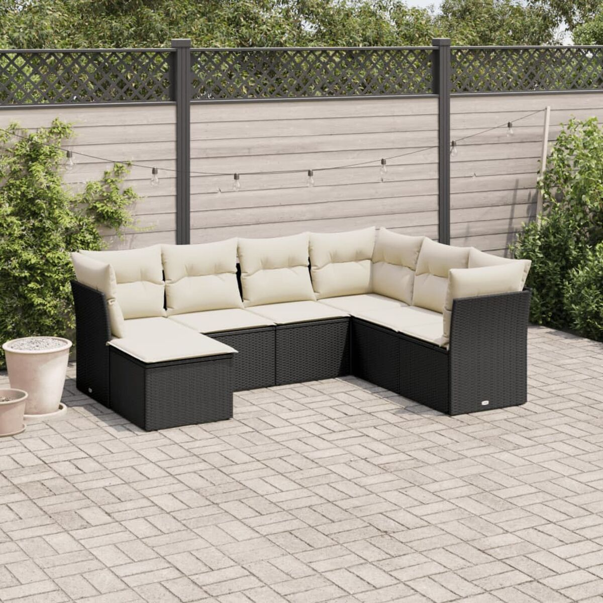 Ein schwarzes Rattan-Ecksofa mit beigen Kissen steht auf einer Backstein-Terrasse. Ein Zaun und Pflanzen sind im Hintergrund.