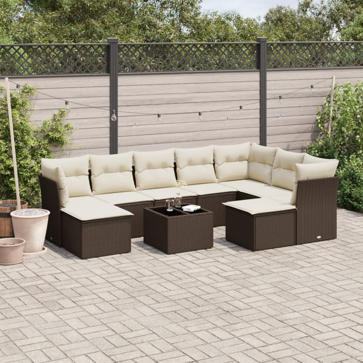 Set divano in rattan marrone con cuscini color crema e tavolino su patio in mattoni, con recinzione in legno.